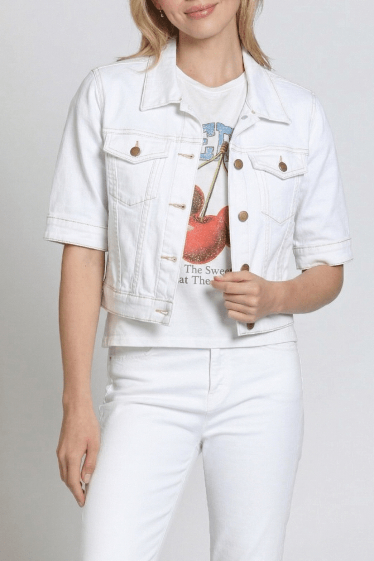 Person wearing a white denim jacket over a graphic t-shirt and white pants on a plain background