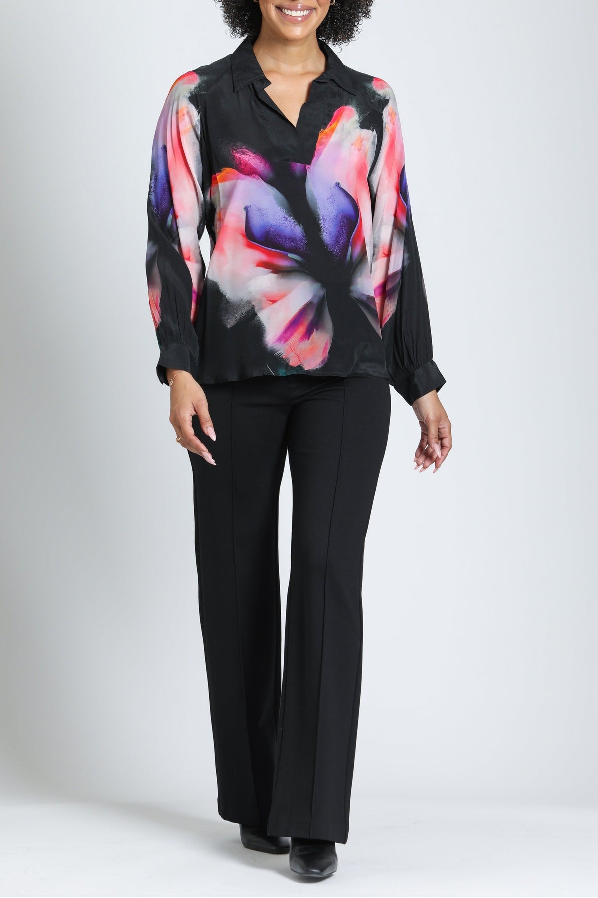 A woman modeling a black long-sleeve blouse with a vibrant pink, purple, and orange abstract floral print, paired with black pants, against a light gray studio background.