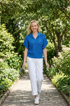 Woman in a blue shirt and white pants walking through a garden path with greenery on either side.