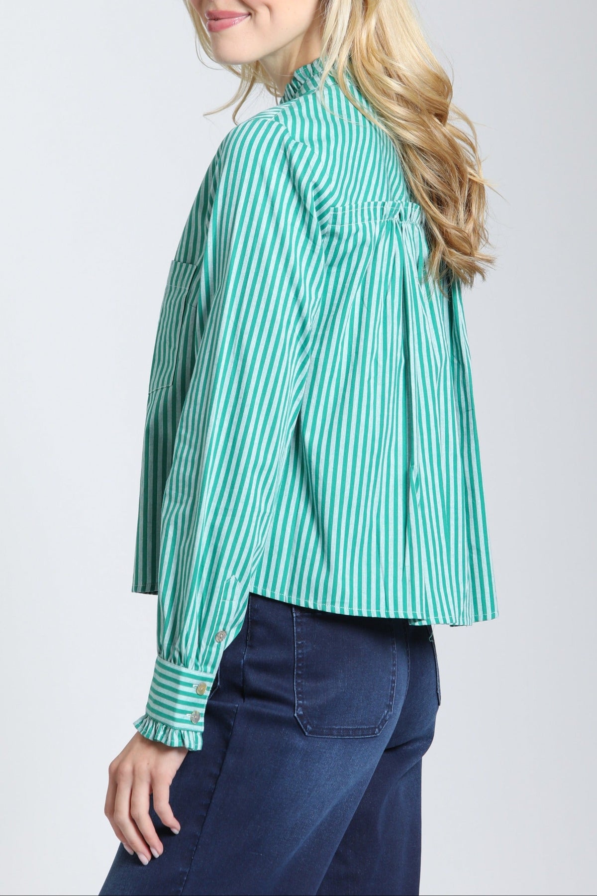 A woman with blonde hair is shown from a side-back angle wearing a mint green and white vertically striped long-sleeve blouse with dark blue jeans, set against a plain light gray studio background.
