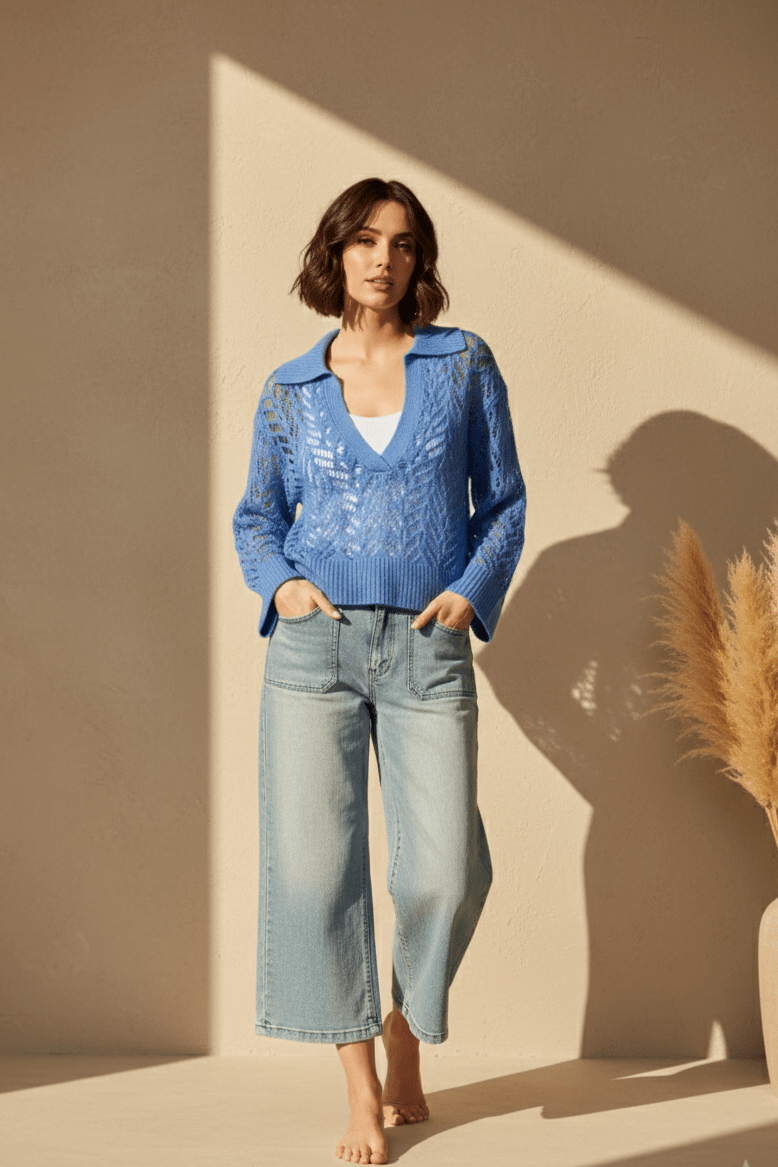 Woman wearing a blue sweater and light blue jeans standing against a beige wall with a plant to the side.