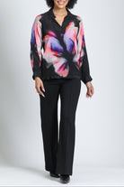 A woman modeling a black long-sleeve blouse with a vibrant pink, purple, and orange abstract floral print, paired with black pants, against a light gray studio background.