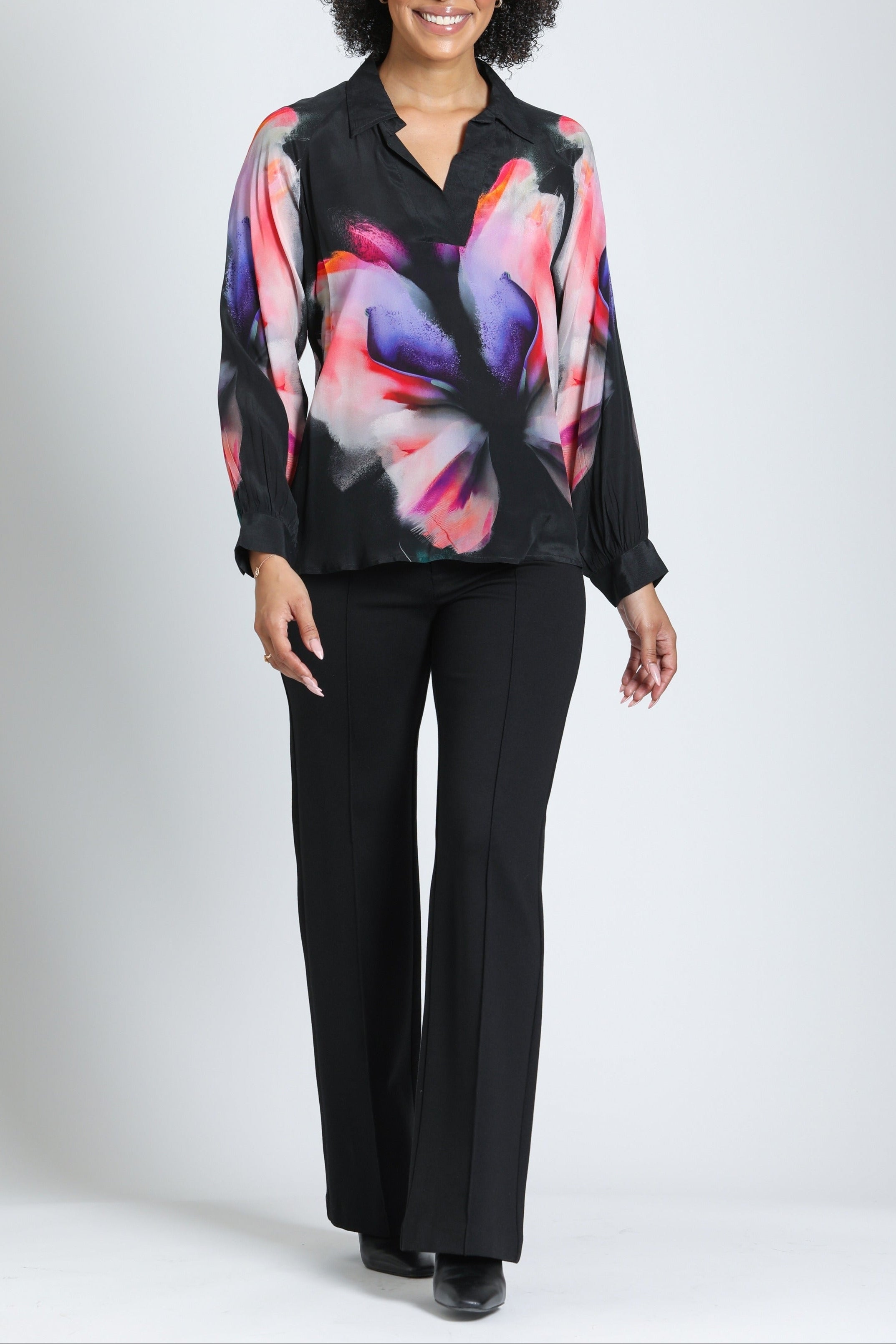 A woman modeling a black long-sleeve blouse with a vibrant pink, purple, and orange abstract floral print, paired with black pants, against a light gray studio background.