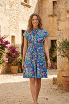 Woman wearing a floral dress standing in an outdoor setting with stone walls and potted plants.