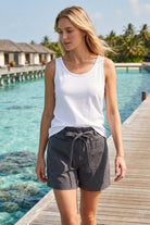 Woman in a white tank top and gray shorts standing on a wooden dock by a pool with a tropical setting.